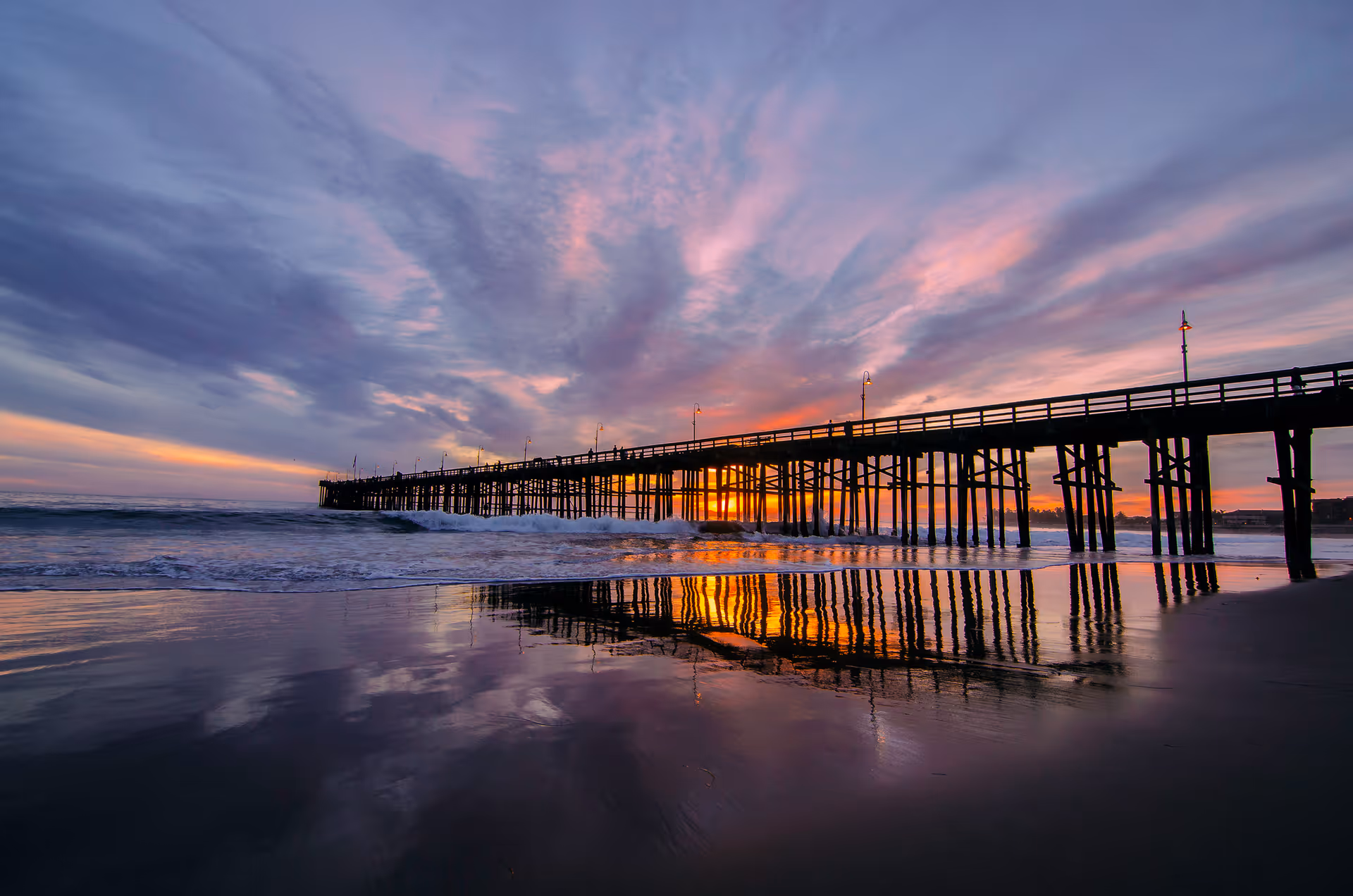 Wooden pier stretching over the ocean at sunset with colorful clouds and reflections on the wet sand.