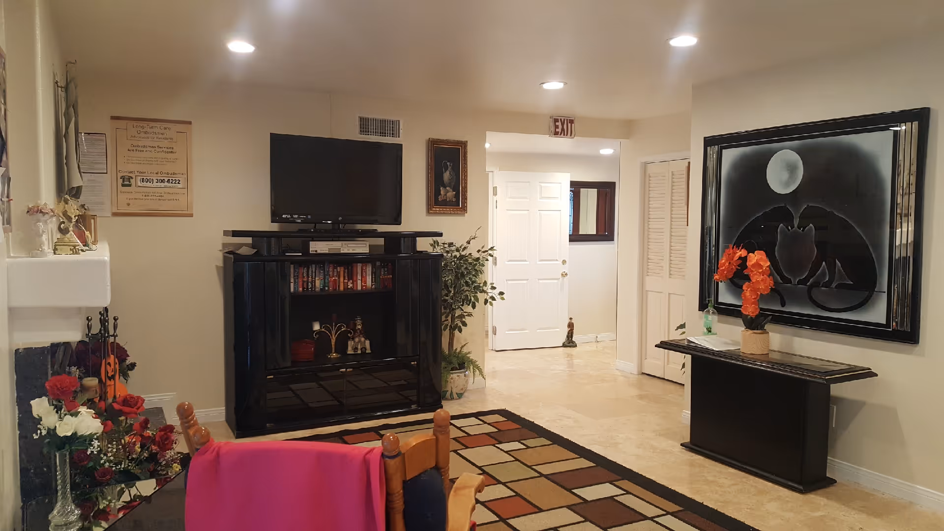Interior view of a senior living facility common area with a TV on a black cabinet, a colorful patterned rug, a wooden chair with a pink cloth draped over it, a fireplace with floral decorations, a large framed artwork of cats under a moon on the wall, and an exit door in the background.