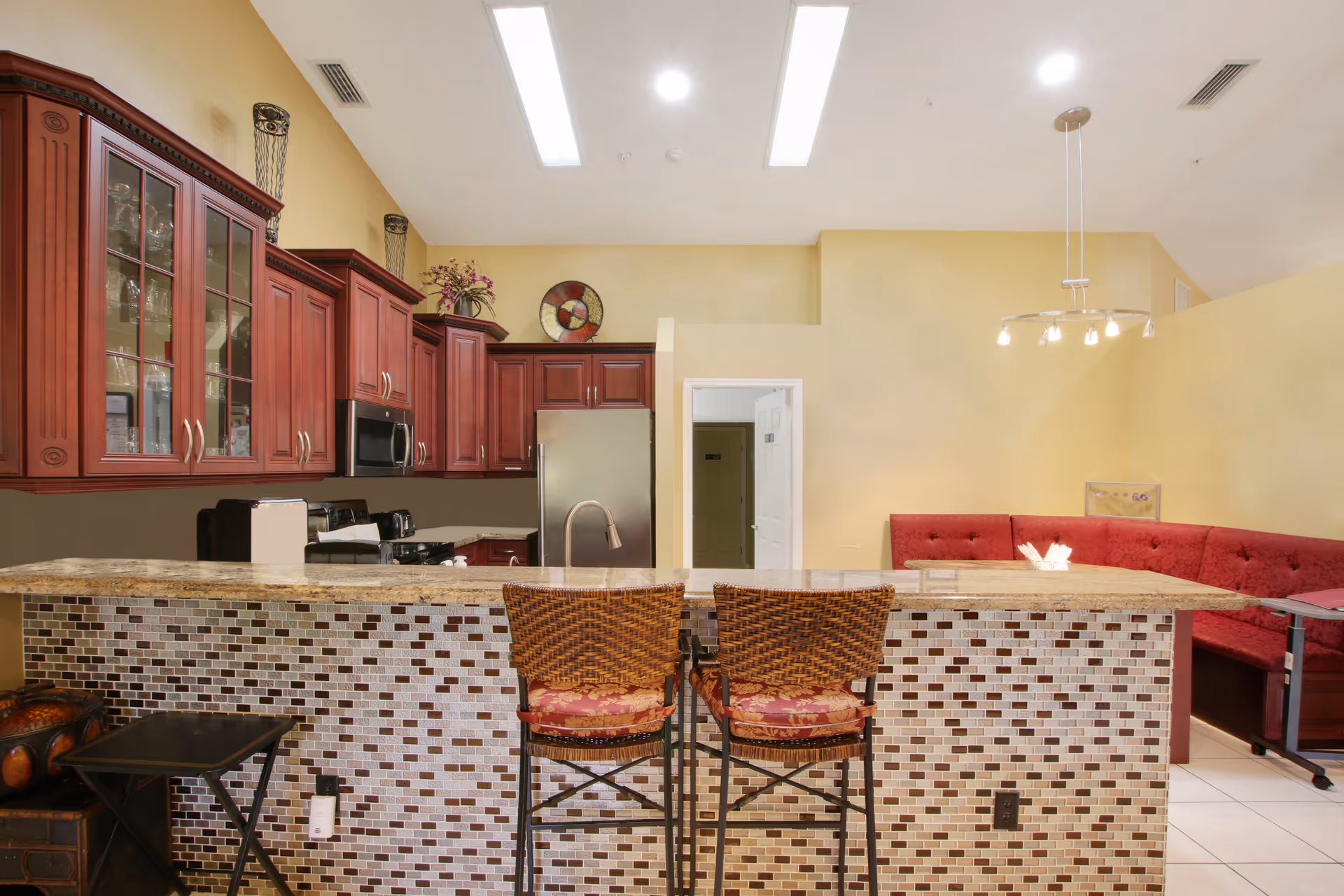 Interior view of a kitchen and dining area in an assisted living facility. The kitchen features dark wood cabinets, a stainless steel refrigerator, microwave, and stove. A tiled breakfast bar with two wicker chairs is in the foreground. In the background, there is a red cushioned bench seating area with a modern light fixture above.