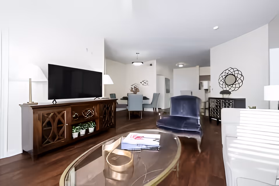 A modern living room with a wooden TV stand holding a flat-screen television, a glass coffee table with a book on it, a blue velvet chair, and a white sofa. In the background, there is a dining area with a table and chairs, decorative wall art, and a console table with a round mirror above it. The room has wooden flooring and white walls.