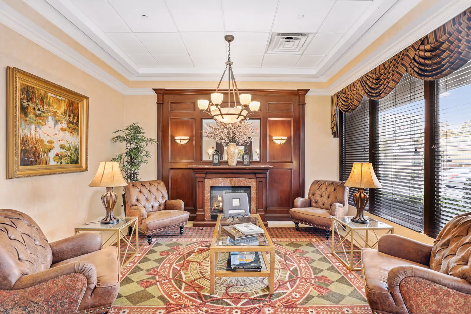 A cozy living room with four brown leather armchairs arranged around a glass coffee table on a patterned rug. The room features a wooden fireplace with a mirror and decorative vases above it, two table lamps on side tables, a large window with blinds and draped curtains, a potted plant, and a framed painting on the wall.