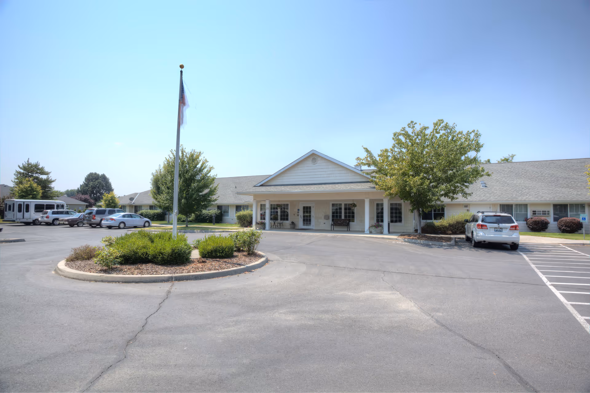 Front exterior view of a single-story senior living facility building with a covered entrance, a flagpole with an American flag, several parked cars, and landscaped greenery under a clear blue sky.