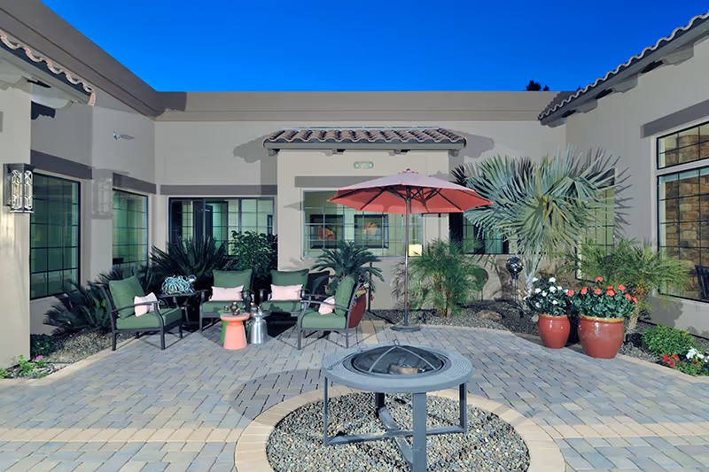 Outdoor courtyard with patio seating, a red umbrella, potted plants and a central fire pit in front of a beige building.