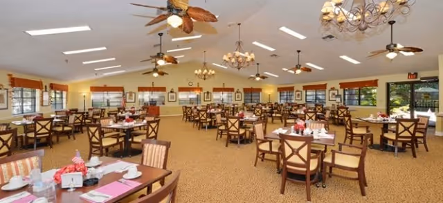 A spacious dining room with multiple wooden tables and chairs arranged neatly. Each table is set with plates, cups, napkins, and small flower centerpieces. The room has large windows with orange valances, ceiling fans with leaf-shaped blades, and elegant chandeliers hanging from a high ceiling with skylights. The carpet is patterned in warm tones, and the overall atmosphere is bright and welcoming.