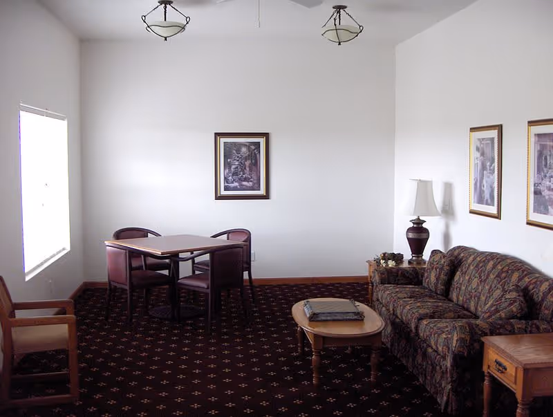 A small living room area with a patterned sofa, wooden coffee table, side table with a lamp, and a dining table with four chairs. The room has white walls, three framed pictures, and two ceiling light fixtures. There is a window on the left side letting in natural light.