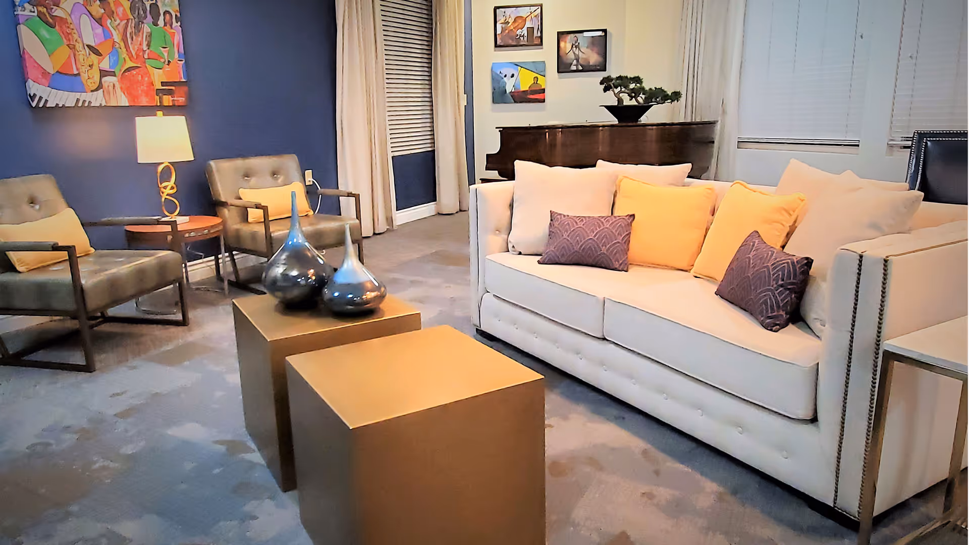 A cozy living room with a white sofa adorned with yellow and purple pillows, two modern armchairs with yellow cushions, a pair of gold square coffee tables with decorative vases, a lamp on a side table, colorful artwork on the walls, and a piano in the background.