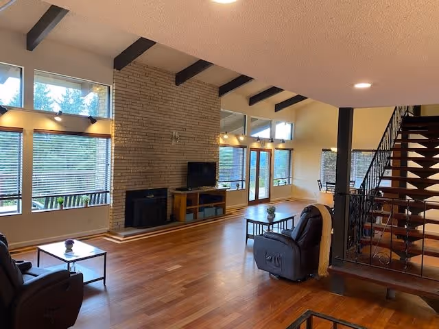 Spacious living room with wooden floors, a brick fireplace, a TV on a wooden stand, large windows with blinds, two recliner chairs, a coffee table, and a staircase with metal railing leading to an upper floor.