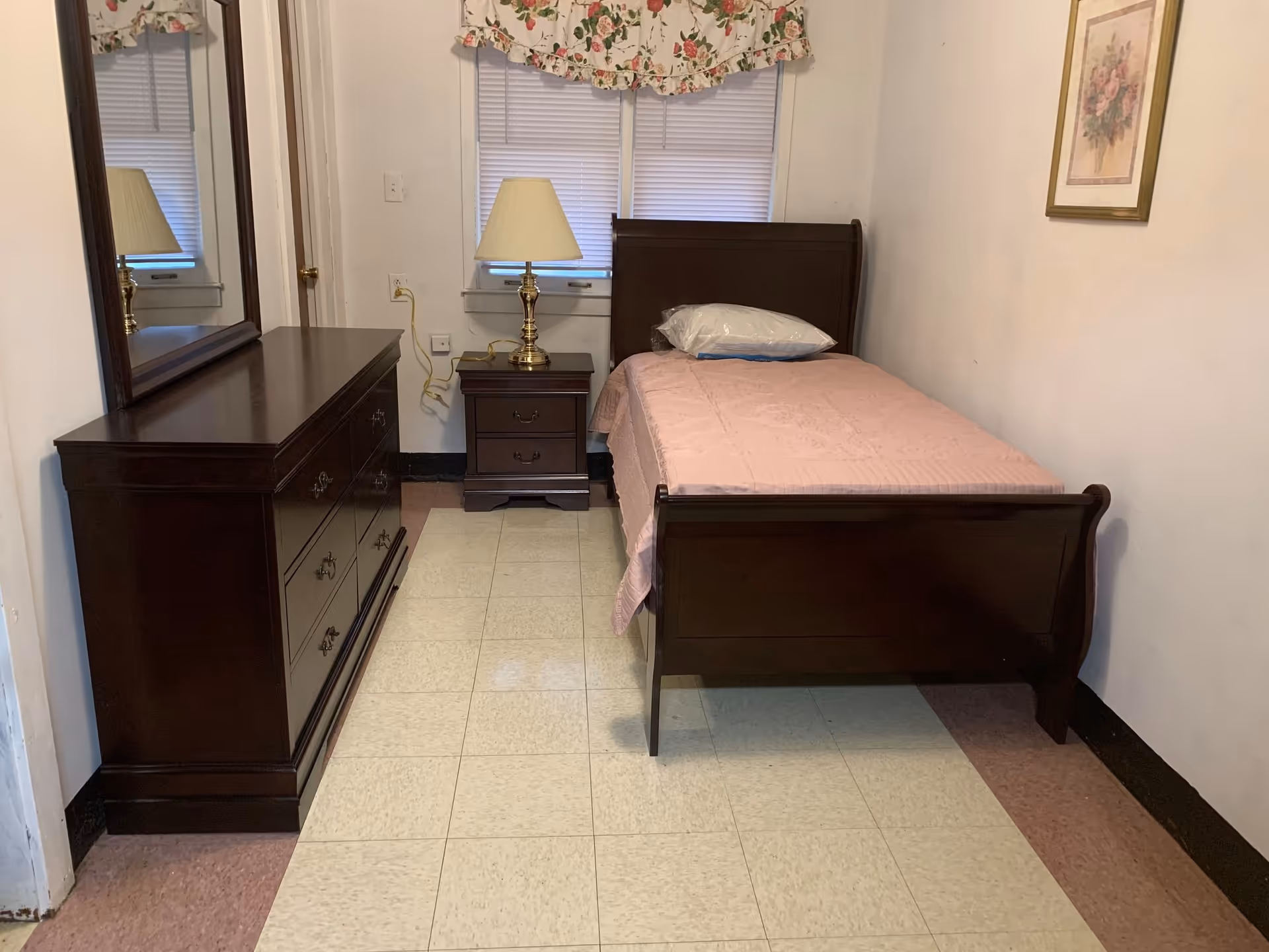 A small bedroom with a single dark wooden bed covered with a pink bedspread and a pillow wrapped in plastic. Next to the bed is a matching dark wooden nightstand with a brass lamp. Across from the bed is a dark wooden dresser with a large mirror. The room has tiled flooring and a window with closed blinds and a floral valance. A framed floral picture hangs on the wall.