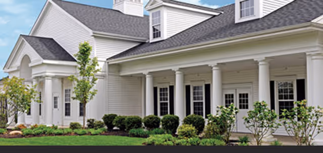 Exterior view of a white building with columns and a well-maintained garden with shrubs and small trees in front under a blue sky.