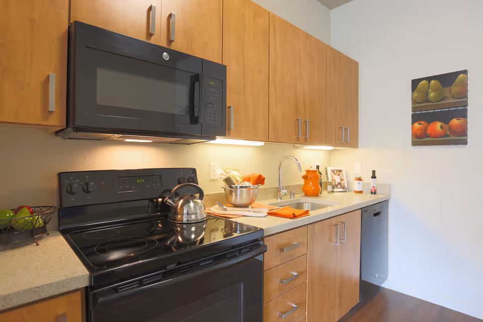 Modern kitchen with wooden cabinets, a black electric stove with a stainless steel kettle, a black microwave above the stove, a countertop with a sink, orange kitchen towels, a bowl with vegetables, and a painting of fruit on the wall.