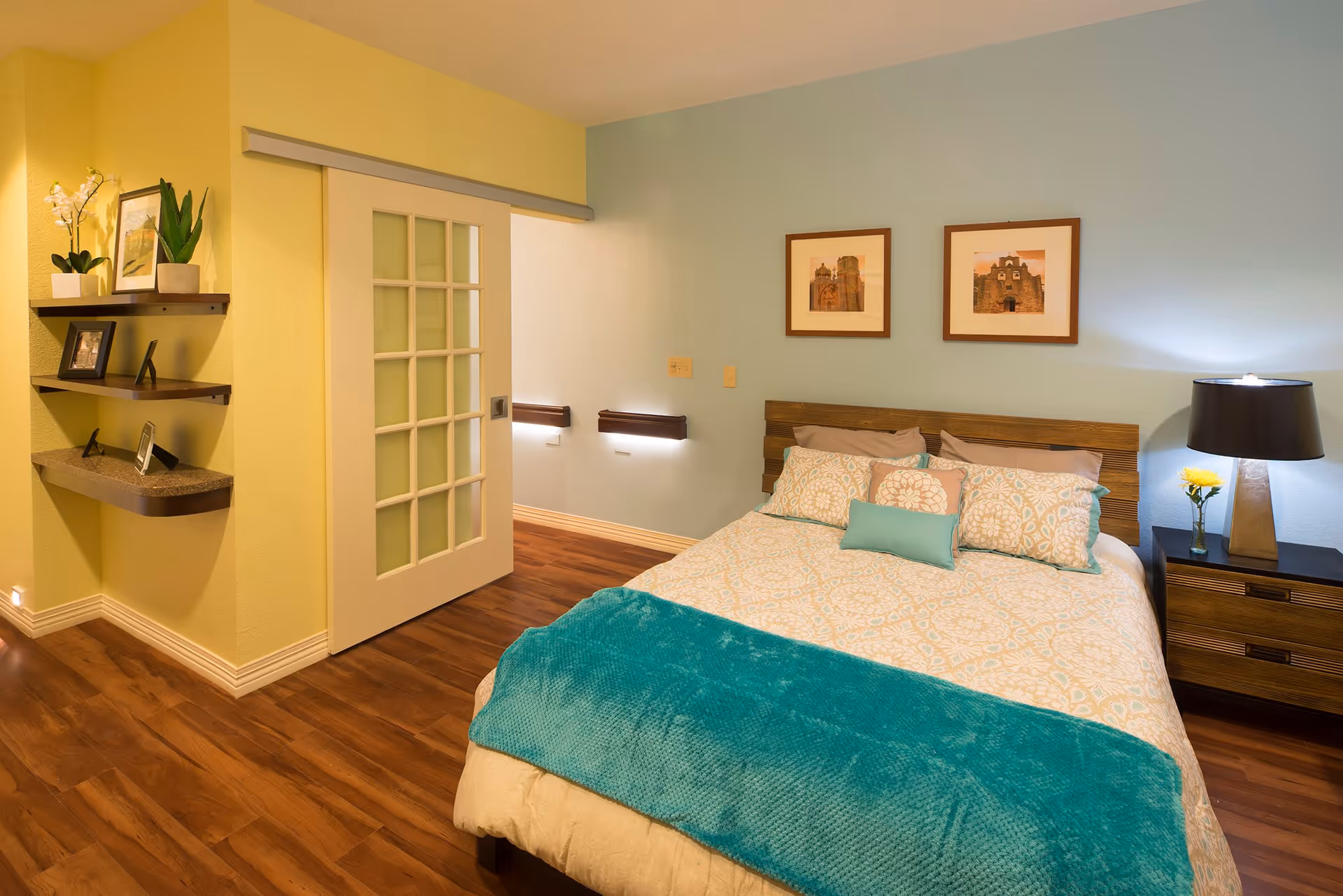 A cozy bedroom with a wooden bed frame, a bed made with patterned bedding and multiple pillows, and a teal throw blanket. There is a wooden nightstand with a lamp and a yellow flower in a vase. The walls are painted light blue and yellow, with two framed pictures hanging above the bed. A sliding door with glass panels leads to another room, and there are floating shelves with plants and picture frames on the yellow wall.