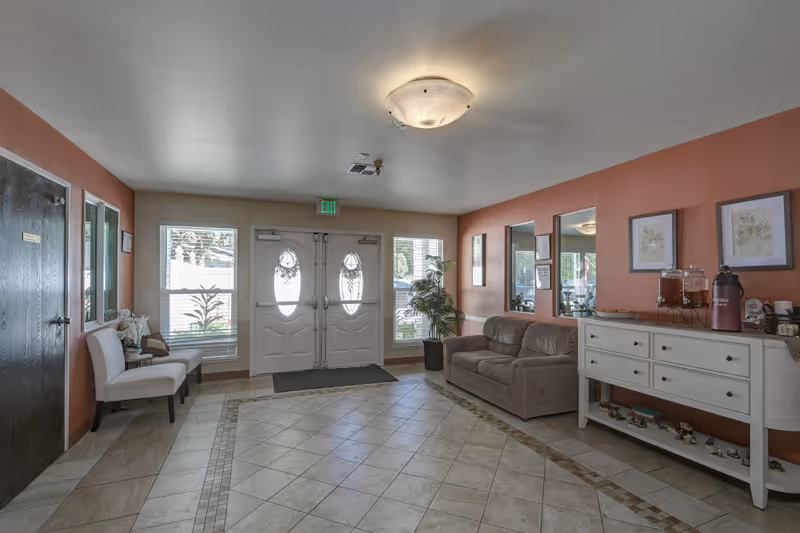 A bright and welcoming assisted living facility entrance area with double doors featuring oval glass panels. The room has tiled flooring with a decorative border, a brown couch, a white sideboard with decorative items, two white chairs, framed artwork on the walls, and a potted plant near the door.