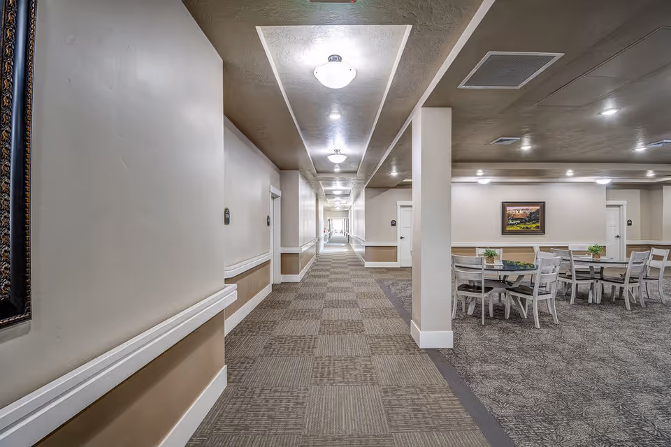 A long, well-lit interior corridor of a senior living facility with handrails along the walls and a seating/dining area to the right.