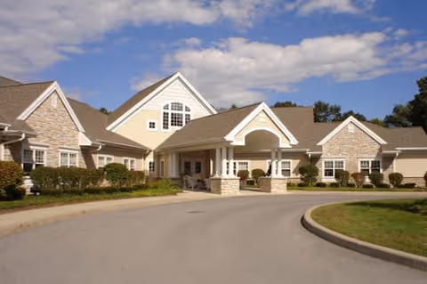 Front exterior view of a senior living facility building with a covered entrance, stone and siding facade, and a curved driveway under a partly cloudy sky.