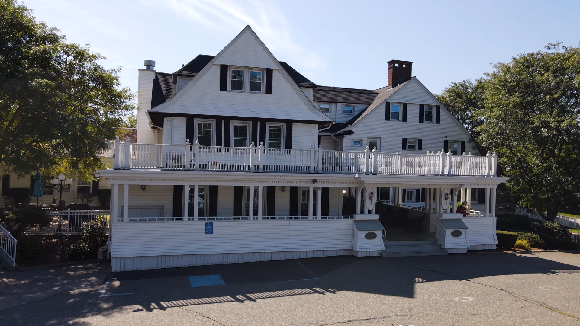 Front exterior of a large white multi-story building with a wraparound porch, upper balcony, and surrounding trees.