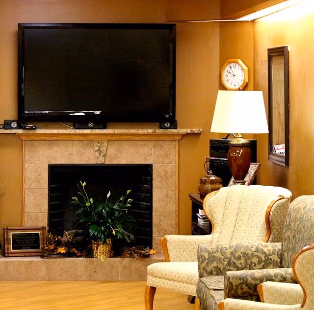 A cozy living room area featuring a mounted flat-screen TV above a beige tiled fireplace with a green potted plant inside. To the right, there is a wooden side table with a brown lamp, a clock on the wall, and a mirror. Two upholstered armchairs with patterned cushions are positioned in front of the fireplace.