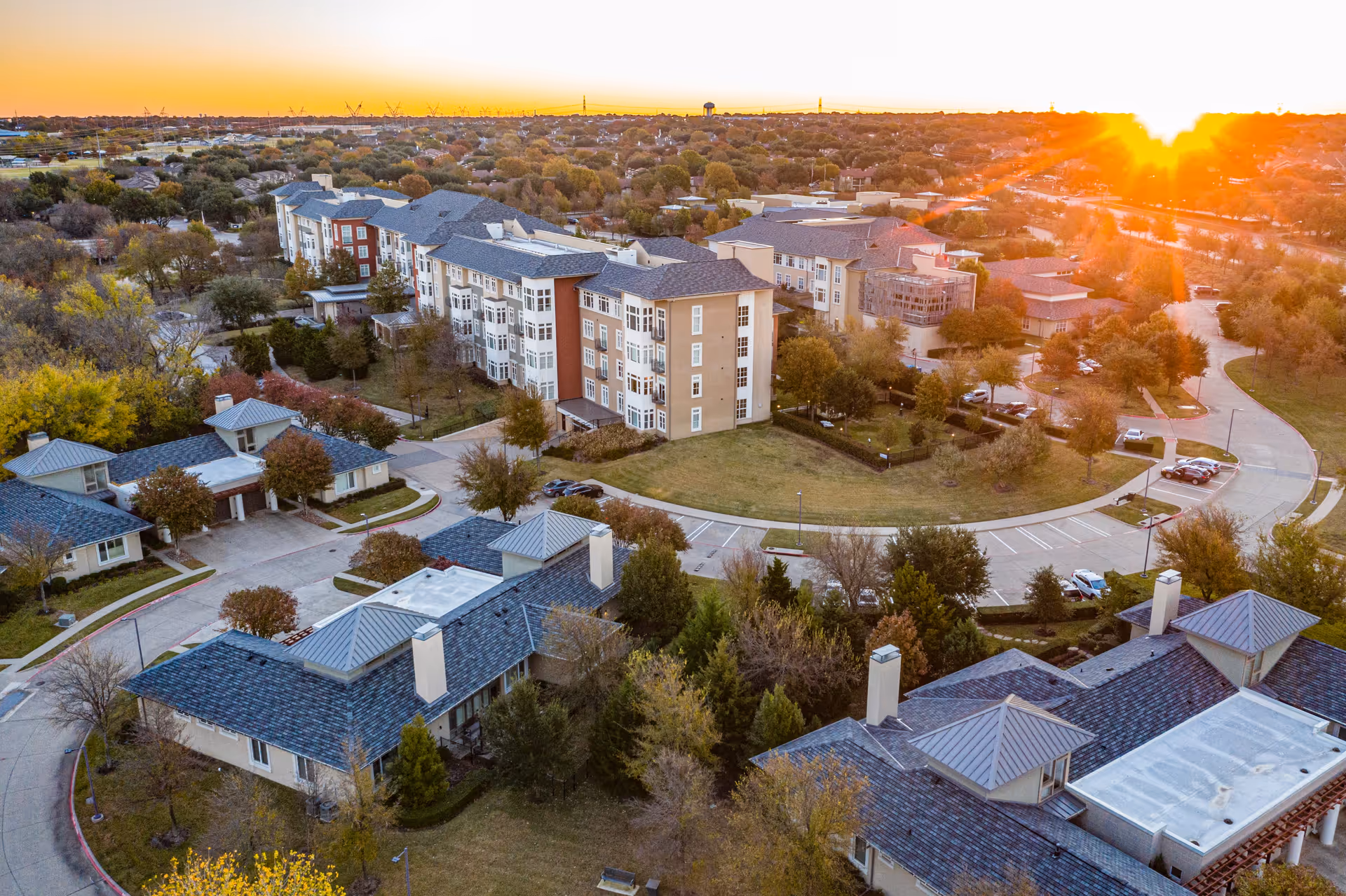 Aerial view of The Legacy Willow Bend campus at sunset showing multi-story residential buildings, smaller cottages, parking areas, roads, and surrounding trees.
