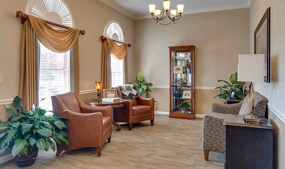 A cozy sitting area in a senior living facility with two brown leather armchairs and a patterned fabric armchair arranged around a small wooden table. The room has two large windows with beige curtains and valances, a wooden display cabinet with glass doors containing decorative items, several green plants, a table lamp, and a chandelier hanging from the ceiling.