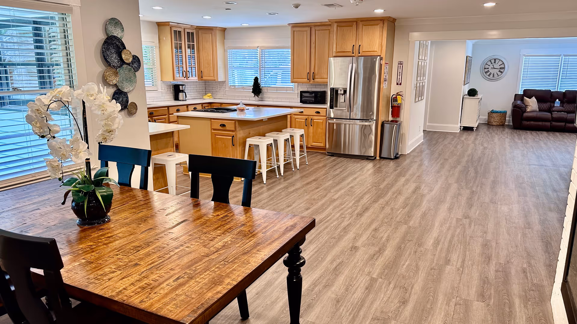 Spacious open kitchen and dining area with wooden cabinets, stainless steel refrigerator, kitchen island with white stools, wooden dining table with black chairs, and a white orchid centerpiece. In the background, a living room area with a brown leather sofa and a wall clock is visible.