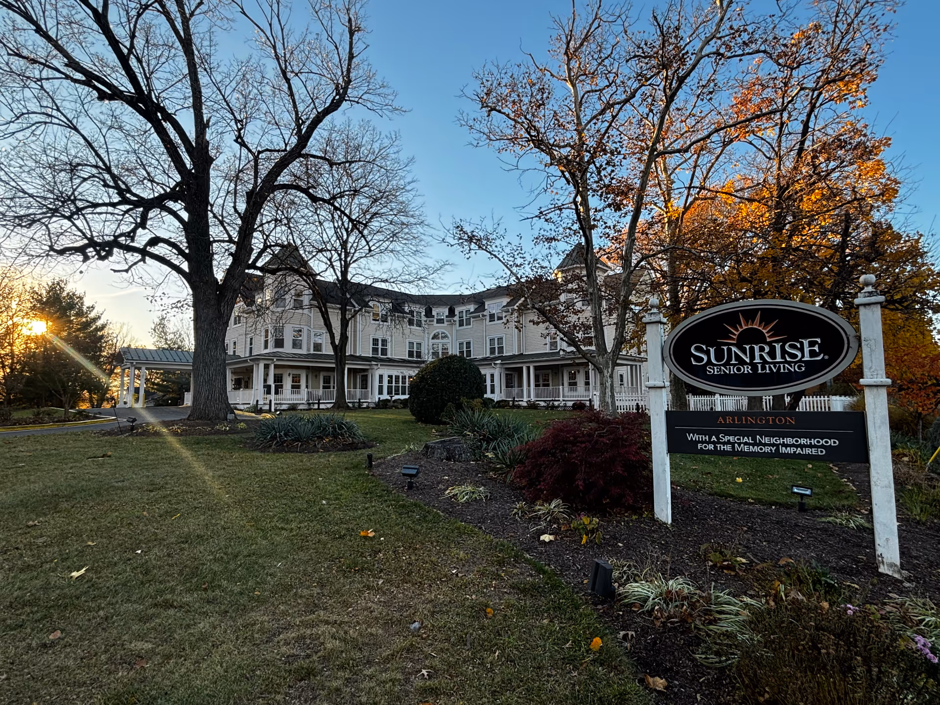 Exterior view of Sunrise Senior Living facility in Arlington during sunset, showing a large multi-story building with a porch, surrounded by trees with autumn foliage and a well-maintained lawn. A sign in the foreground reads 'Sunrise Senior Living Arlington With a Special Neighborhood for the Memory Impaired.'