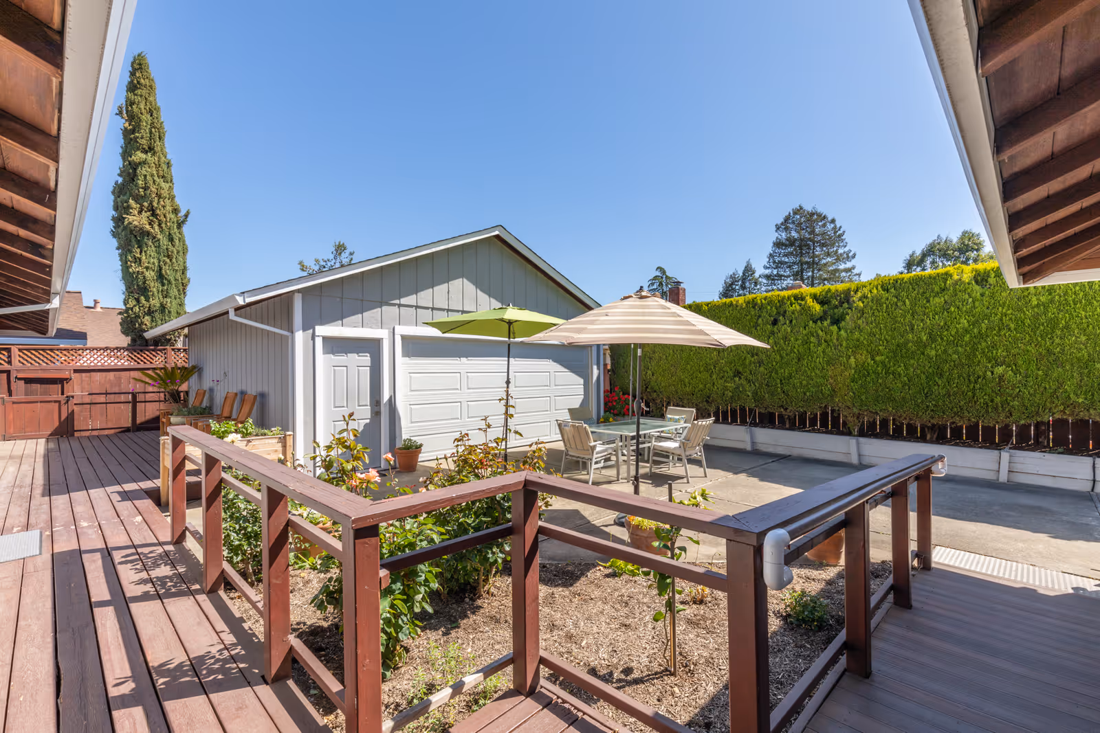 Outdoor patio area with wooden decking and railing, a small garden bed, and a concrete patio with a table and chairs under two umbrellas. A gray garage with a white door and a tall green hedge are visible in the background under a clear blue sky.