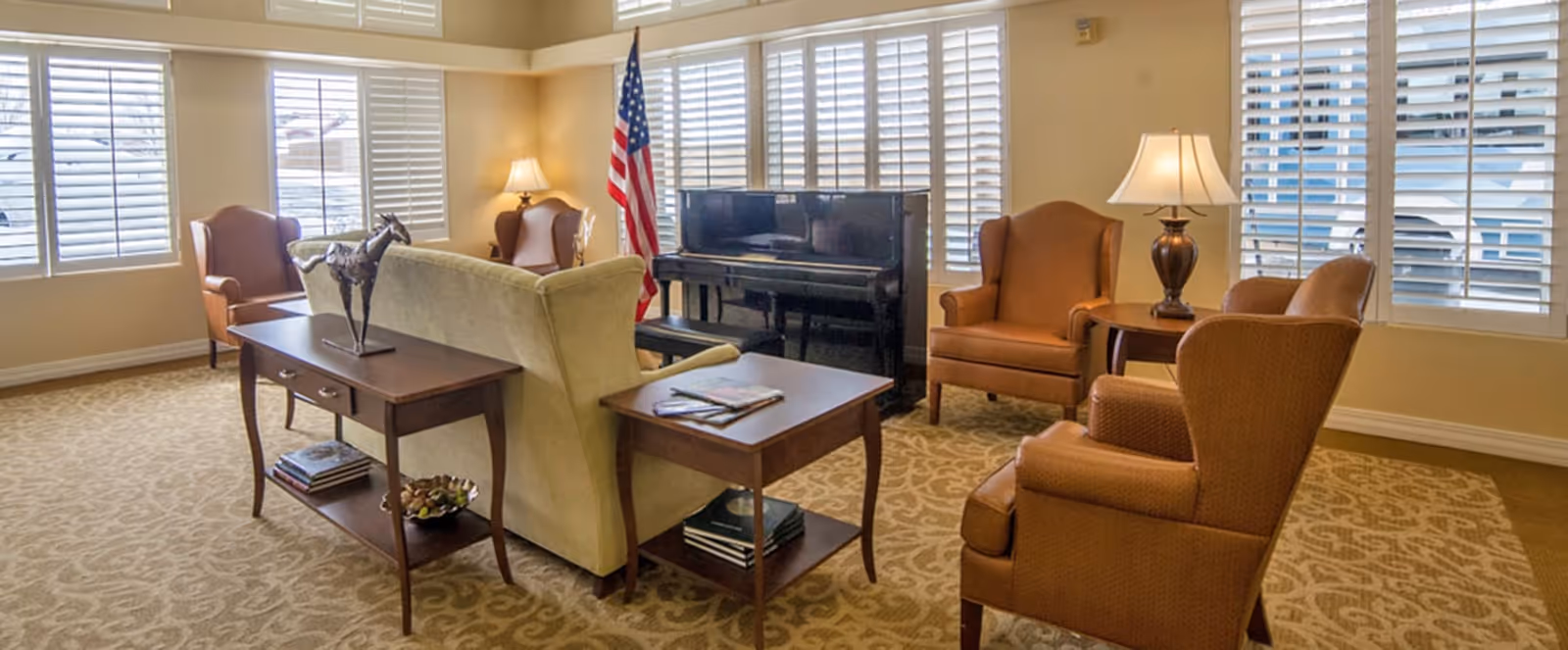 A cozy living room area with beige walls and carpet, featuring a cream-colored sofa, four brown armchairs, two wooden side tables with lamps, a decorative horse sculpture, and a black upright piano near windows with white shutters. An American flag stands in the corner.