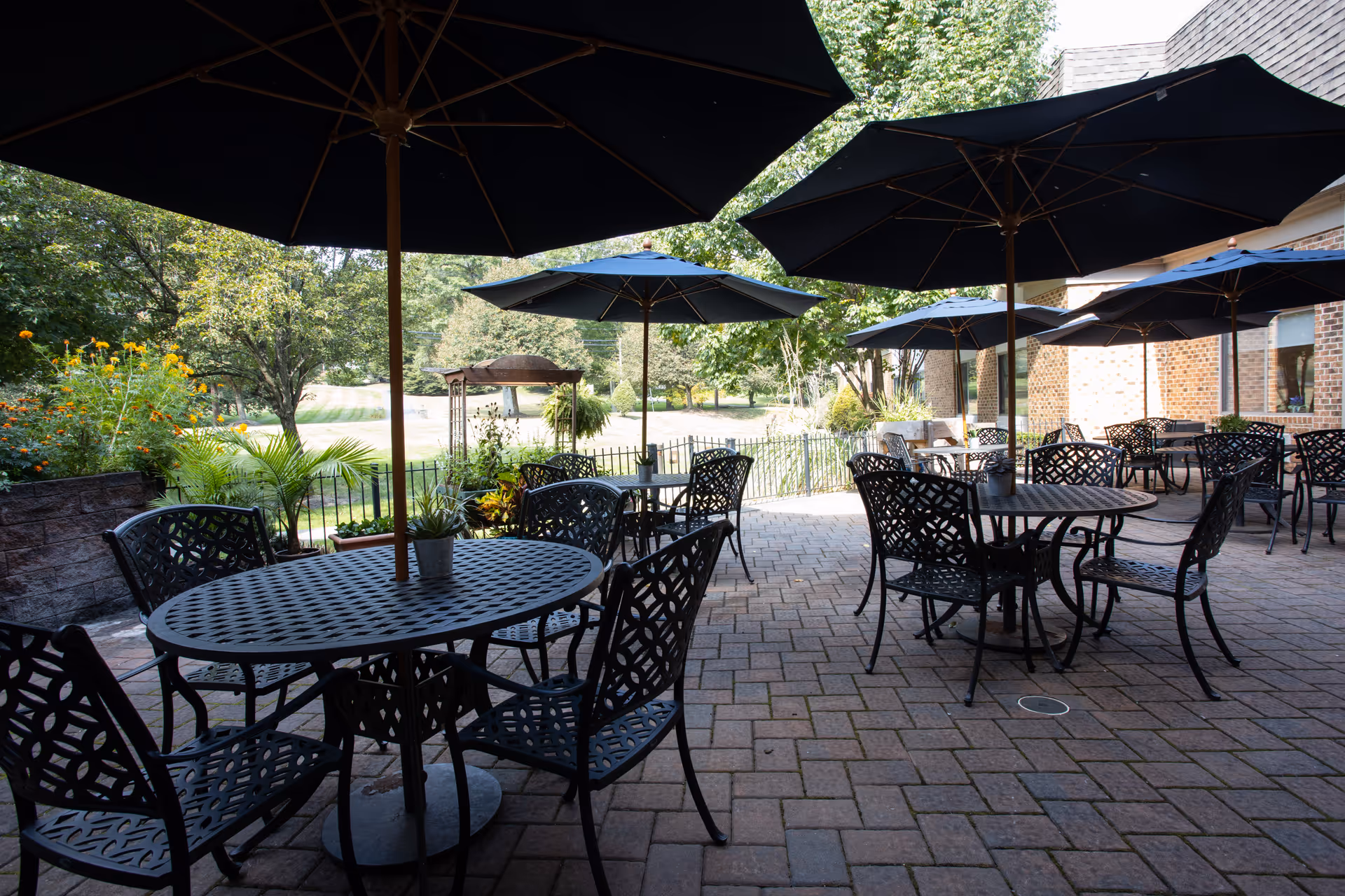 Outdoor patio area with several black metal tables and chairs, each table shaded by a large black umbrella. The patio is paved with brick and surrounded by greenery, including trees and plants, with a brick building visible on the right side.