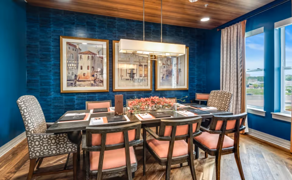 Dining room with a long table surrounded by upholstered chairs, teal walls, framed artwork, and large windows.