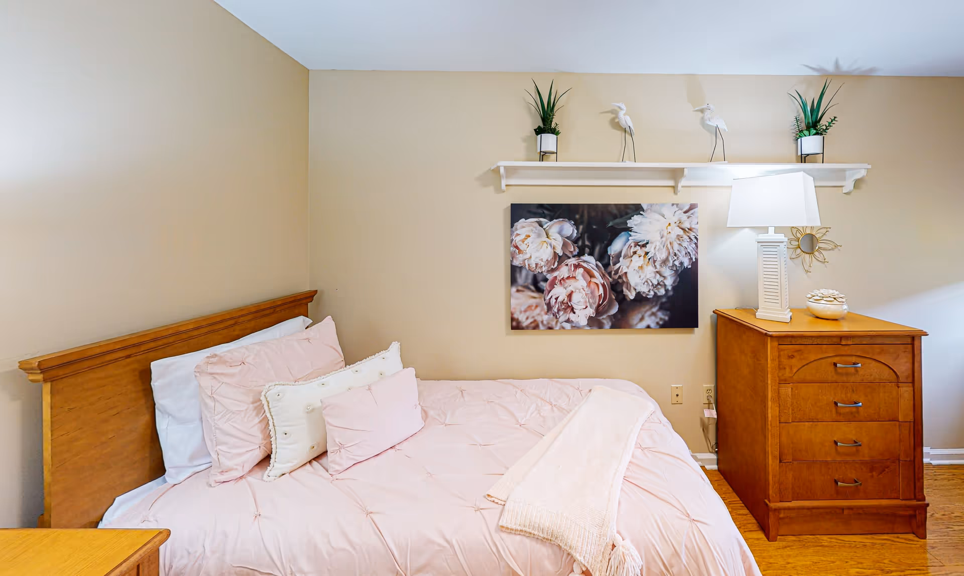 A cozy bedroom with a wooden bed frame and a neatly made bed featuring light pink bedding and multiple pillows. Above the bed, there is a white shelf with two small potted plants and two decorative bird figurines. On the wall below the shelf, there is a large floral artwork. To the right of the bed, there is a wooden dresser with a white table lamp and a decorative bowl on top. The room has beige walls and wooden flooring.