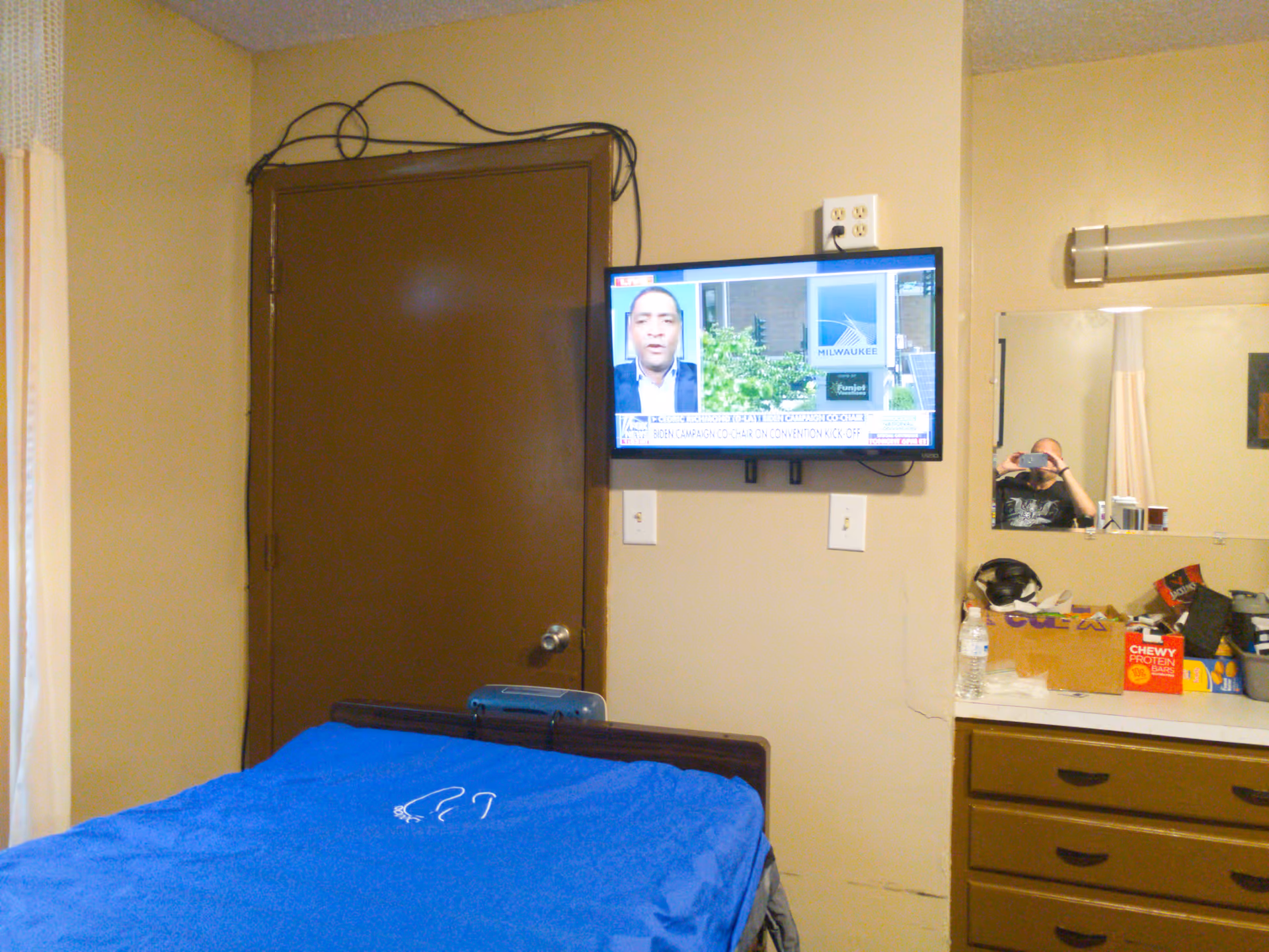 Small patient bedroom with a blue-covered bed, a wall-mounted TV, a brown door, and a dresser with a mirror and personal items.