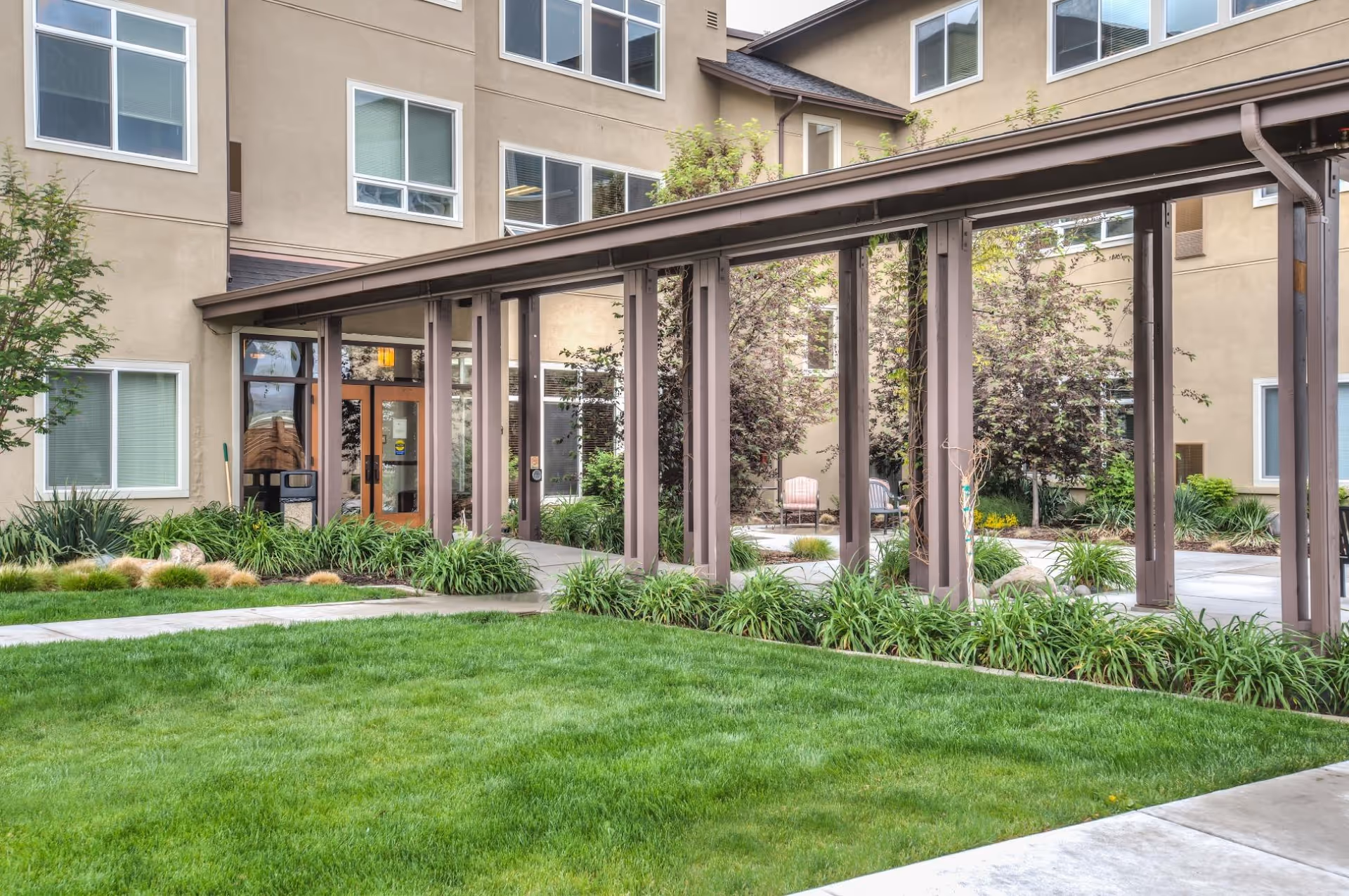 Outdoor courtyard area of The Lodge Assisted Living And Memory Care Community featuring a covered walkway with brown pillars, green grass, landscaped plants, and beige building walls with multiple windows.