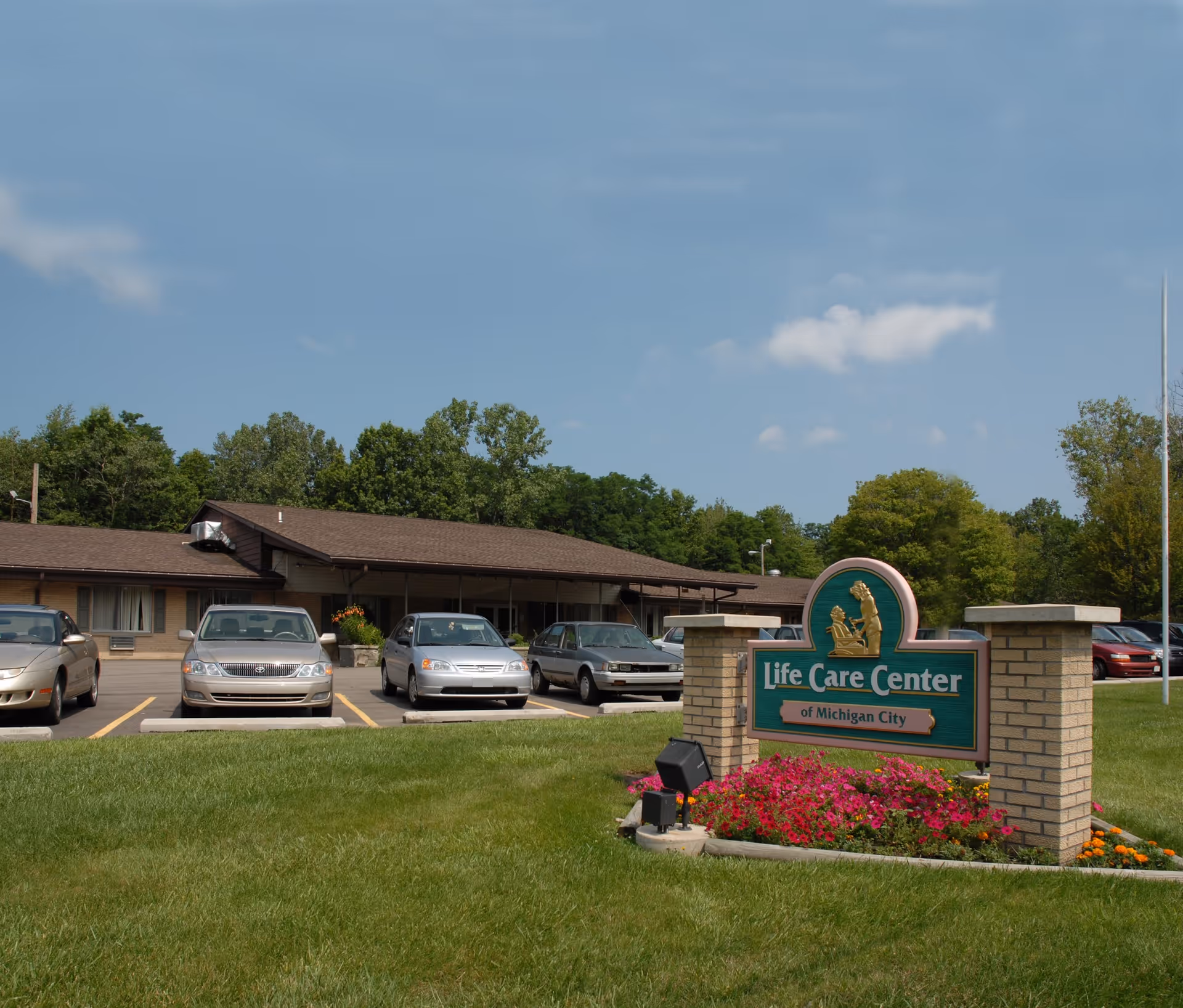 Exterior view of Life Care Center of Michigan City showing a single-story building with a brown roof, several parked cars in front, a green lawn, and a sign with the facility's name surrounded by flowers.