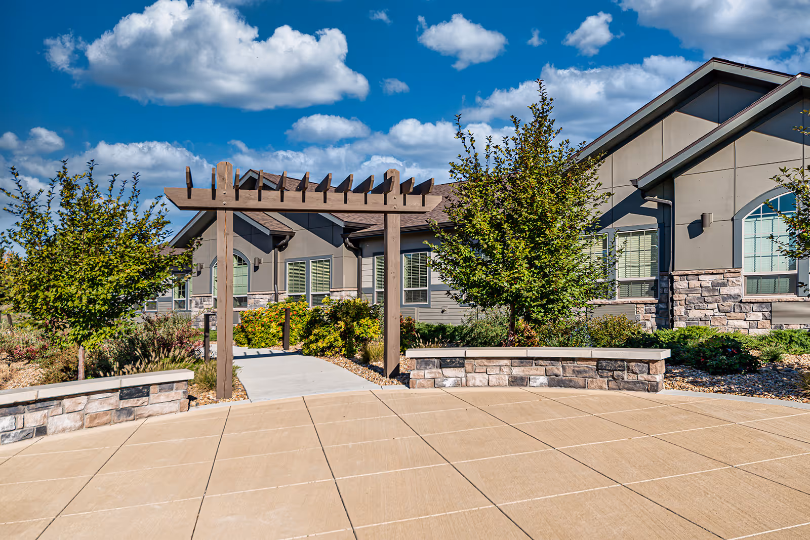 Outdoor view of a senior living facility with a paved patio area, a wooden pergola, landscaped bushes and trees, and a building with stone and siding exterior under a blue sky with scattered clouds.