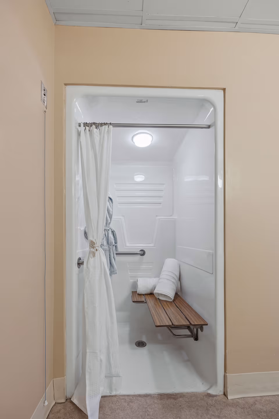 A walk-in shower with a white curtain partially drawn to the side. Inside the shower, there is a wooden fold-down bench with two rolled white towels on it. A grab bar is mounted on the wall, and a bathrobe hangs on a hook. The shower has a ceiling light and a drain on the floor.