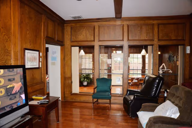 A cozy living room with wooden panel walls and hardwood floors. The room contains a green upholstered chair, a black recliner, and a brown armchair with a white pillow. A flat-screen TV is on a wooden table to the left, and there is a windowed partition with blinds and a view into another room with a table and chairs. Pendant lights hang in the adjacent room.