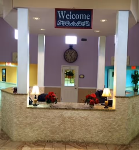 Reception area with a stone-textured counter, two table lamps, red flower arrangements, a wall clock, and a door in the background under a 'Welcome' sign.