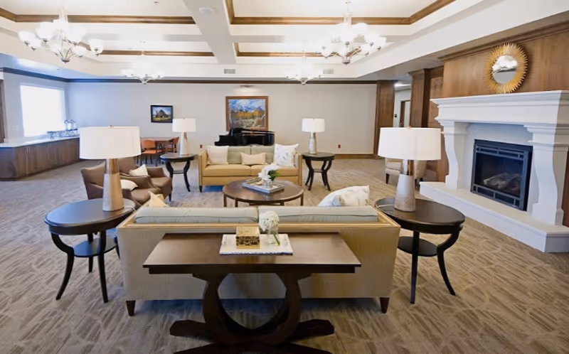 Spacious living room area in an assisted living facility featuring beige sofas with cushions, round wooden coffee tables, side tables with lamps, a white fireplace with a decorative mirror above it, chandeliers on the ceiling, and framed artwork on the walls.