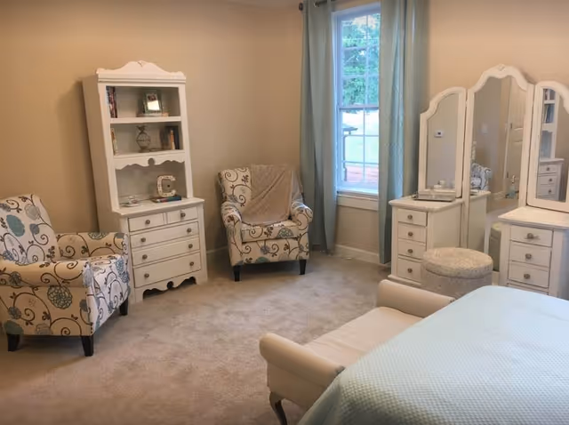 A cozy bedroom featuring two patterned armchairs, a white wooden bookshelf, a window with light blue curtains, a white vanity with three mirrors and drawers, a cushioned stool, and a bed with a light blue blanket.