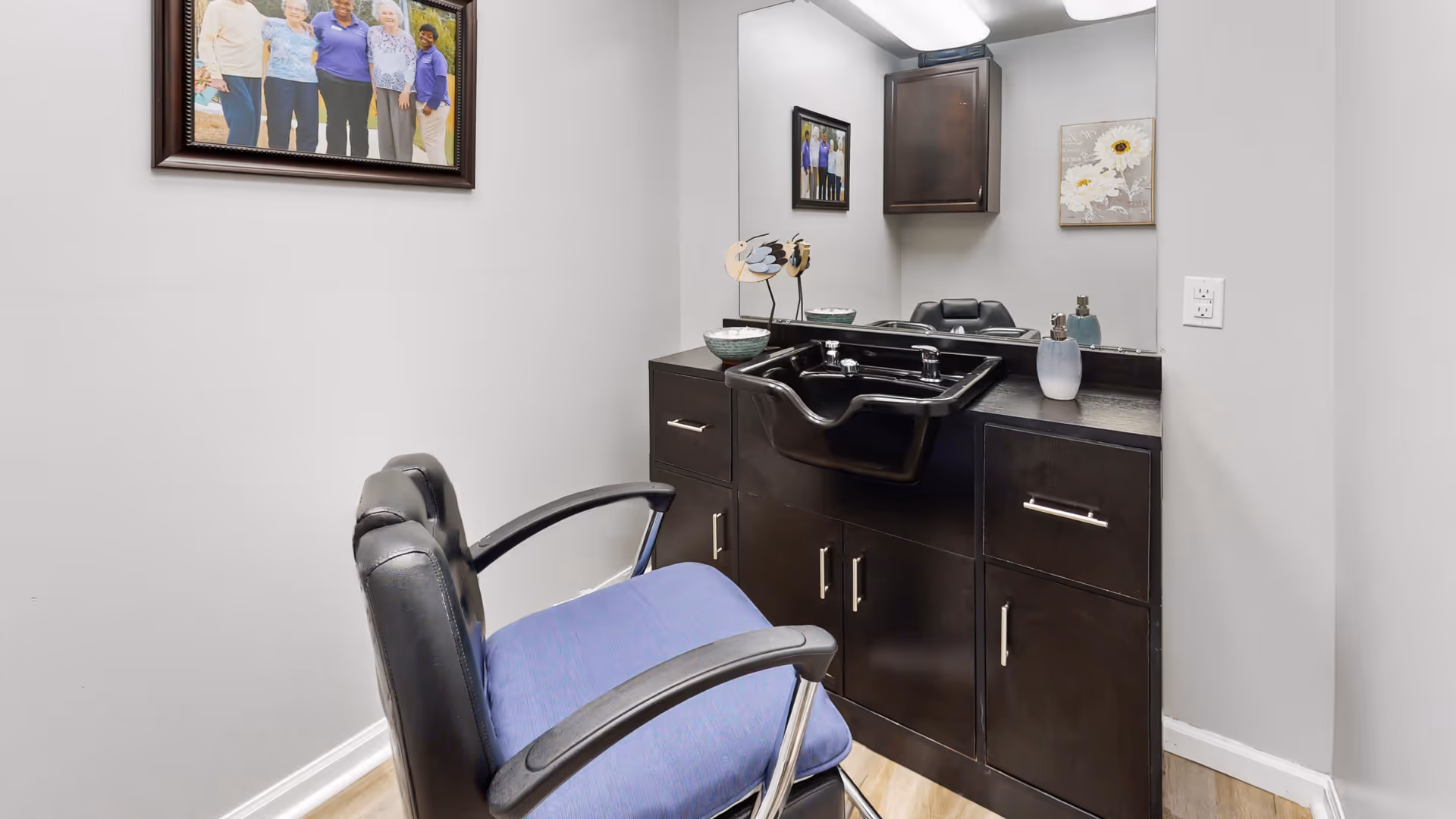 A small salon area with a black salon chair featuring a blue cushion, positioned in front of a black sink and dark wood cabinetry. A large mirror is mounted on the wall above the sink, with a framed photo of a group of elderly people on the adjacent wall and a small decorative flower painting beside the mirror.