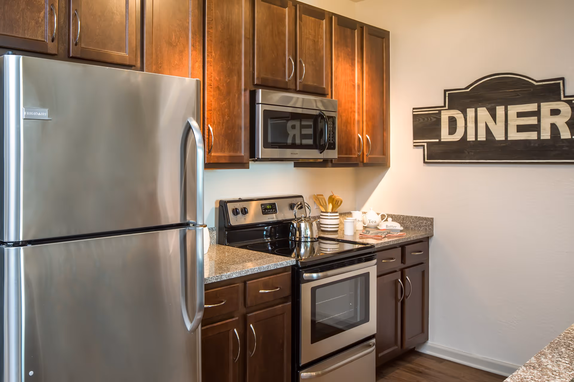A modern kitchen with stainless steel refrigerator, stove, and microwave. The kitchen features dark wooden cabinets and granite countertops. On the right wall, there is a decorative wooden sign that reads 'DINER'. Various kitchen utensils and containers are placed on the countertop.