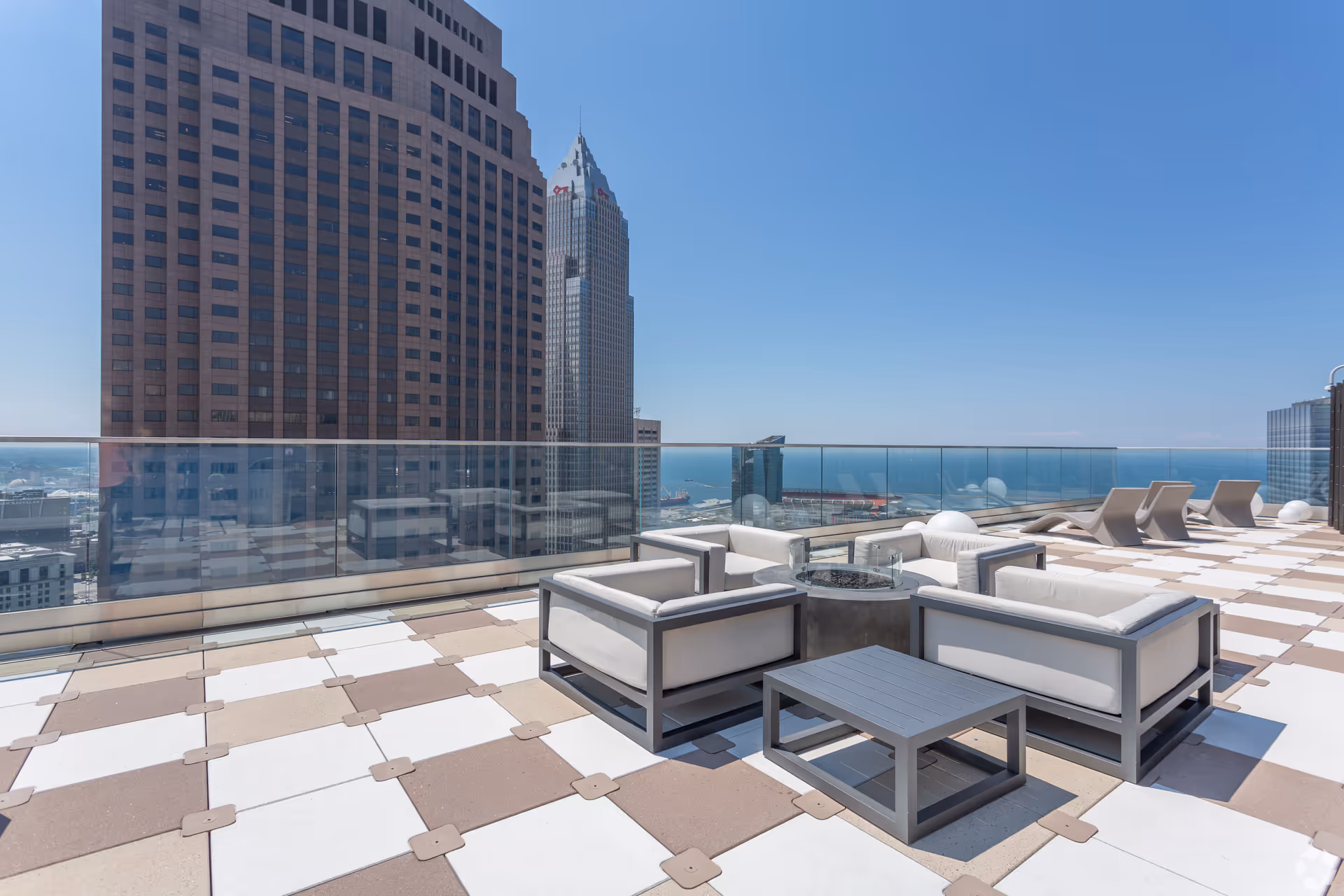 Rooftop patio area with modern outdoor seating including cushioned chairs and a small table, glass railing overlooking tall buildings and a distant view of water under a clear blue sky.