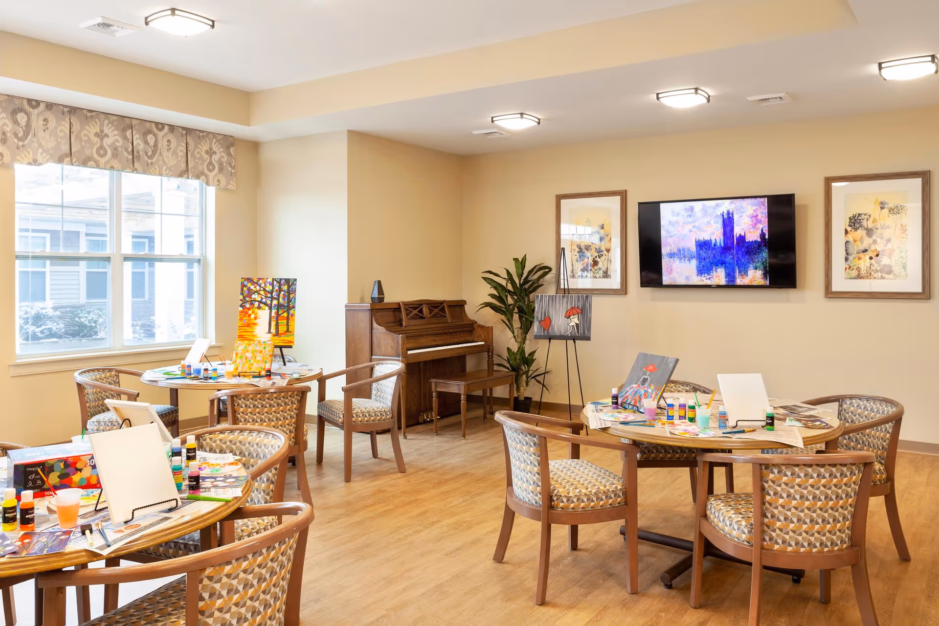 A bright activity room with wooden floors and beige walls featuring several round tables with chairs arranged around them. The tables are set up for painting with canvases, paint bottles, brushes, and palettes. A piano is placed against the far wall next to a tall green plant. Above the piano, a flat-screen TV displays colorful artwork, flanked by two framed pictures. Large windows on the left side allow natural light to fill the room.