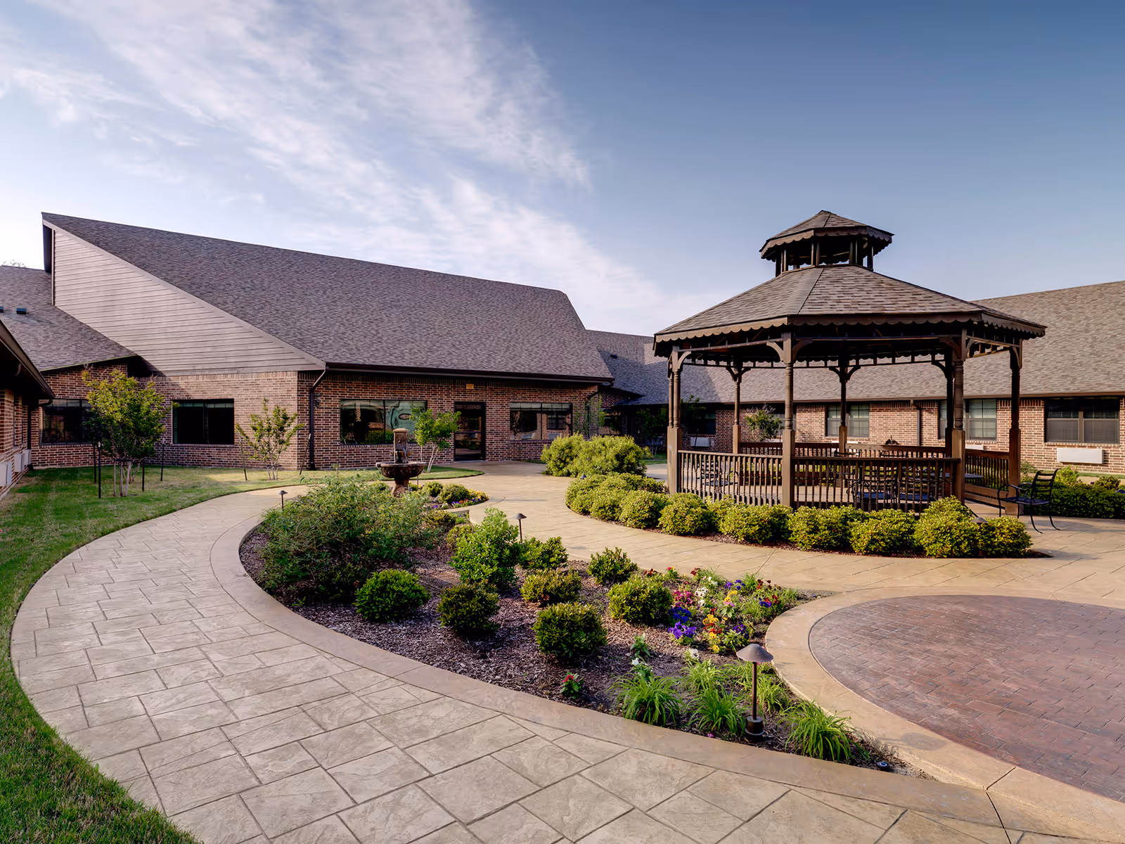 Brick senior living facility courtyard with a wooden gazebo, winding paved paths, a fountain, and landscaped gardens.