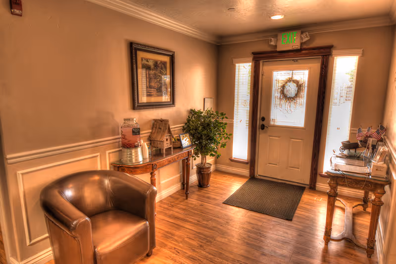 A warmly lit entryway with a wooden floor, a brown leather armchair on the left, a wooden console table with a beverage dispenser and decorative items, a potted plant, and a door with frosted glass panels and a wreath. An exit sign is visible above the door.