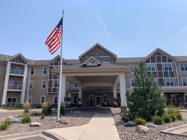 Front exterior view of a multi-story senior living facility named Gable Pines with a covered entrance, an American flag on a flagpole, and landscaped areas with rocks, plants, and small trees under a clear blue sky.