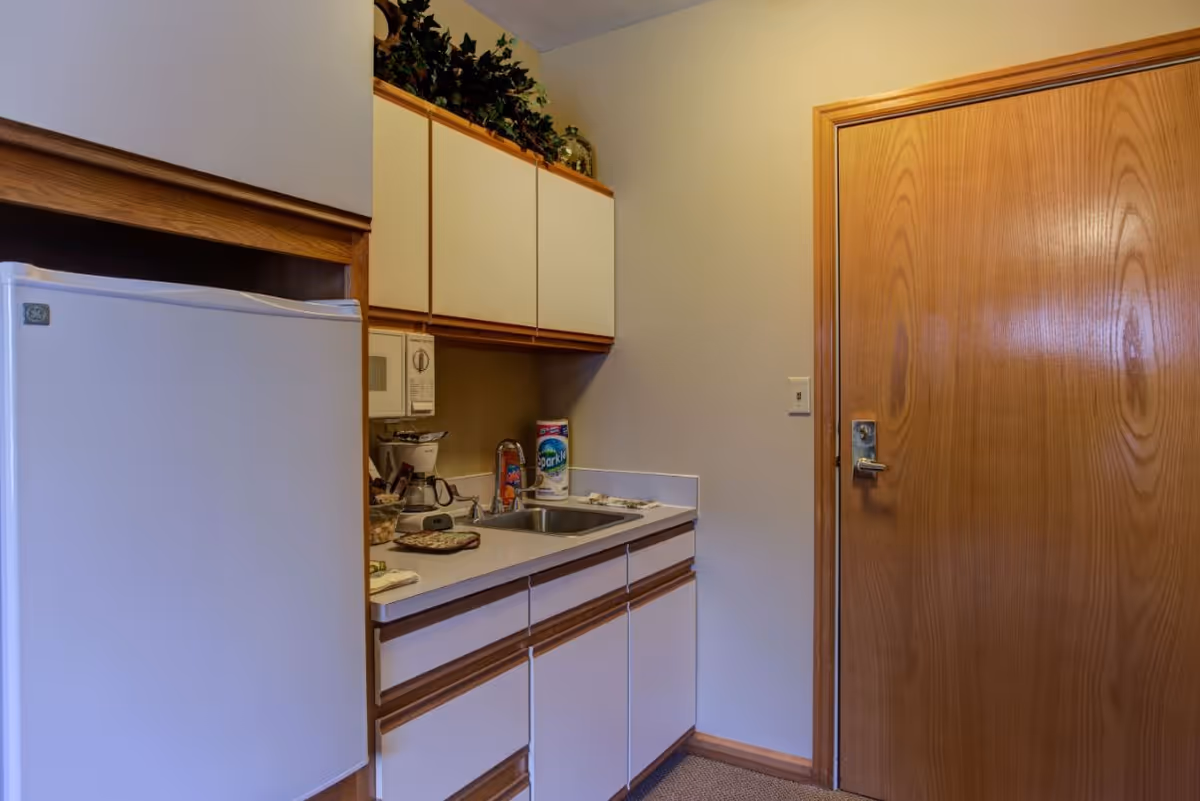 Small kitchenette with a refrigerator, sink, countertop and cabinets next to a wooden door.