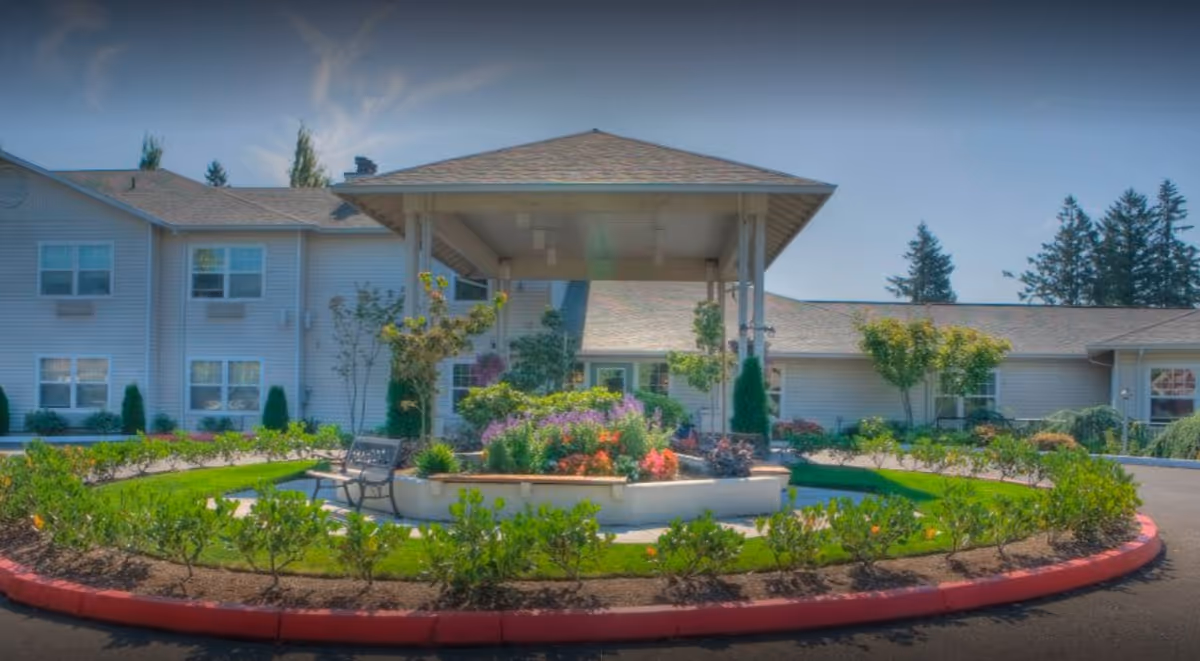 Outdoor circular garden area with a covered gazebo structure in the center, surrounded by colorful flowers, shrubs, and a bench. The background shows a light-colored building with multiple windows and a clear blue sky.