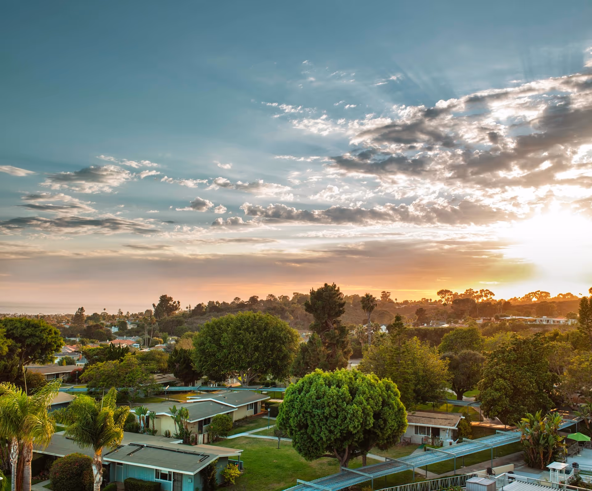 A scenic view of a retirement community with single-story buildings surrounded by lush green trees and lawns under a partly cloudy sky during sunset.