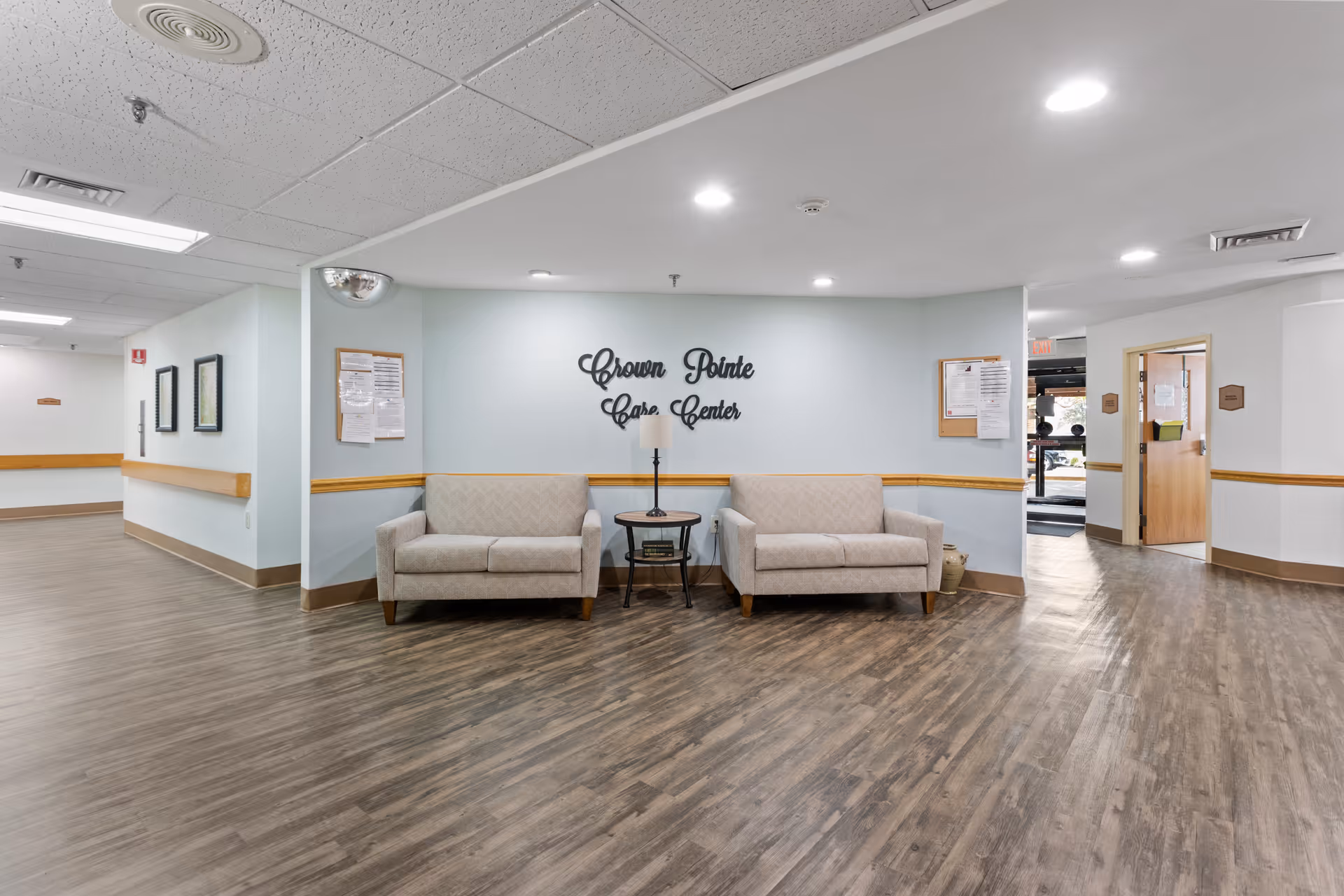 A clean and well-lit hallway area in Crown Pointe Care Center featuring two beige upholstered sofas separated by a small round table with a lamp. The walls are light blue with wooden chair rails, and the floor has wood-patterned vinyl flooring. Bulletin boards with notices are mounted on the walls, and the facility name is displayed in decorative black lettering on the wall behind the sofas.