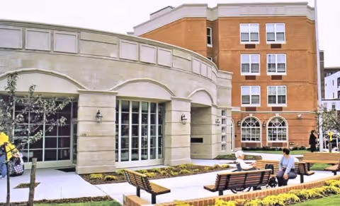 Outdoor courtyard area of a senior living facility with benches, landscaped flower beds, and two people sitting and talking. The building has a brick and stone facade with large windows.